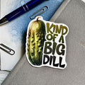 Sticker of a pickle with text 'Kind of a Big Dill' on a notebook with a pen and paper clips.