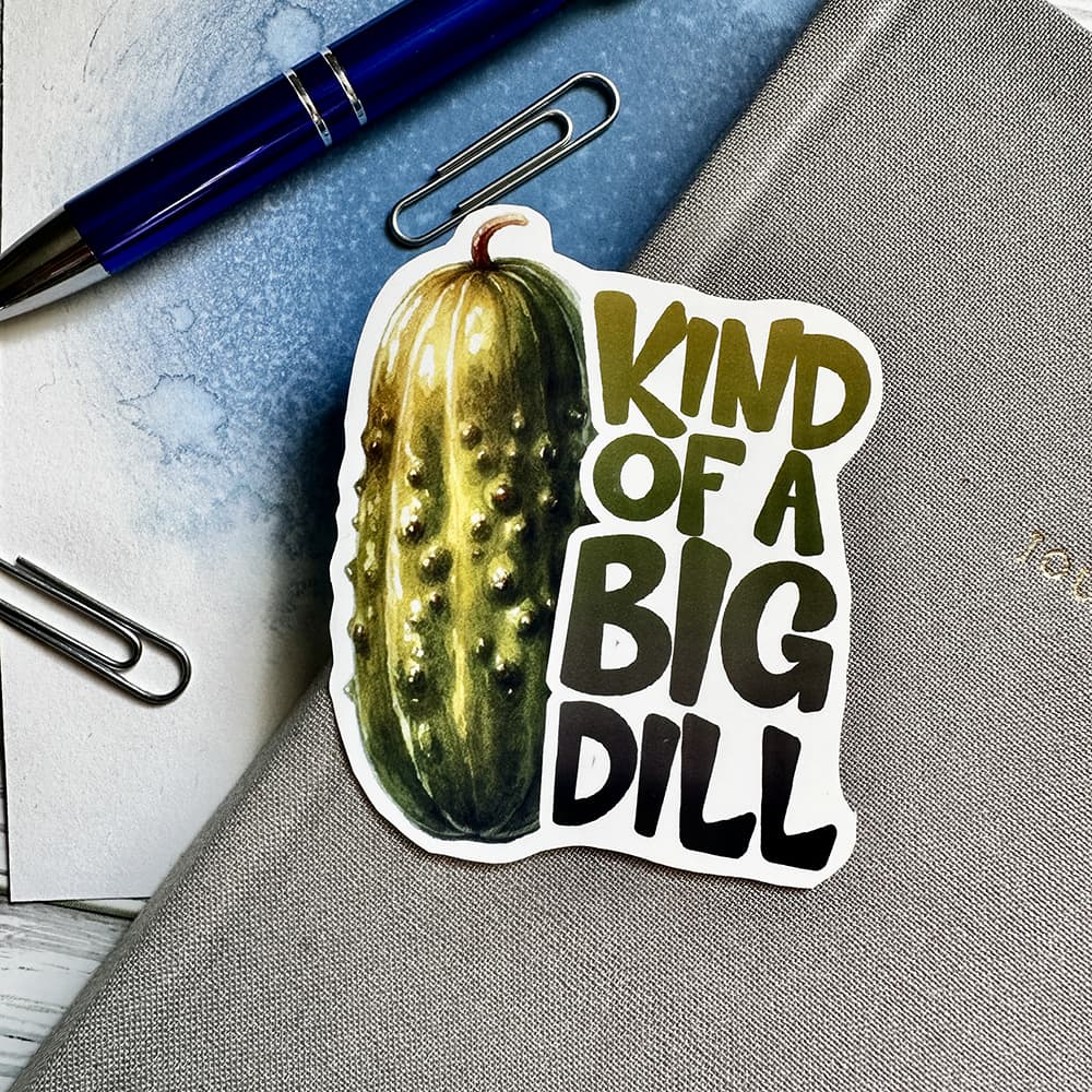Sticker of a pickle with text 'Kind of a Big Dill' on a notebook with a pen and paper clips.