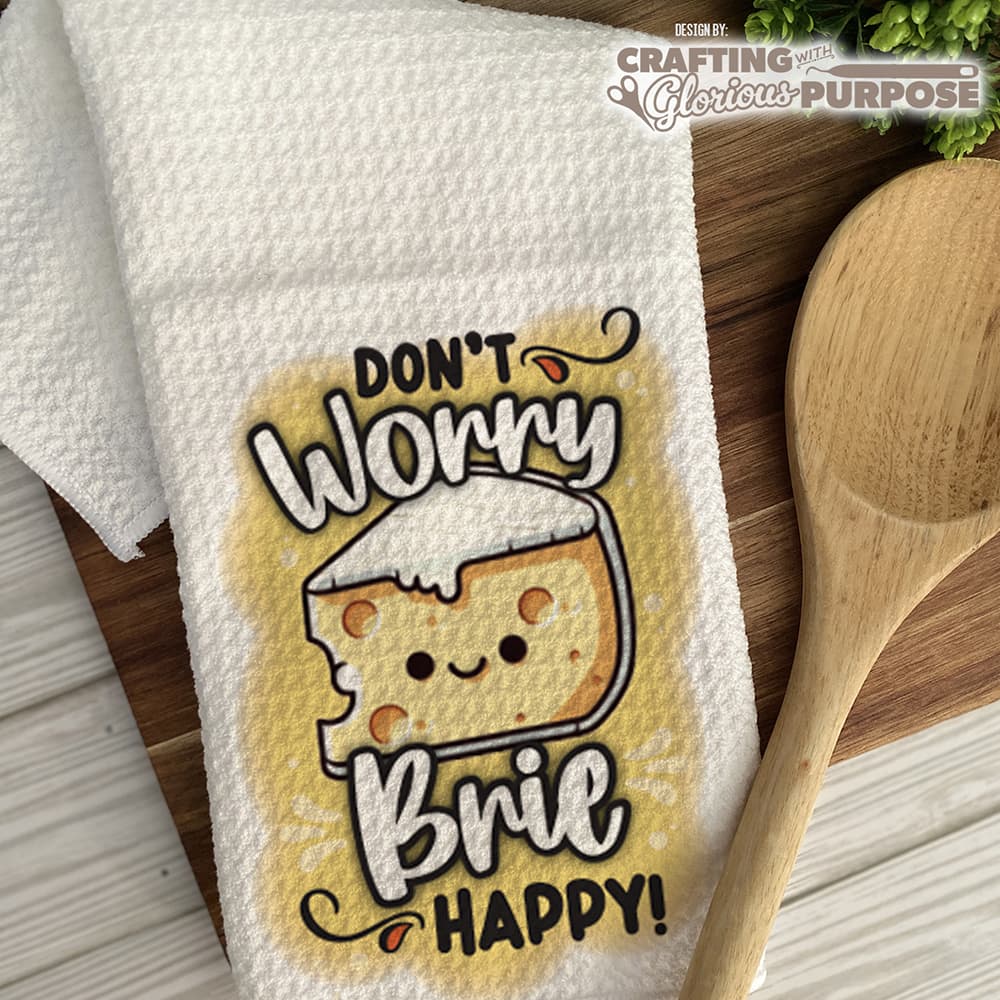 Towel with a cartoon brie cheese character and text 'Don't Worry Brie Happy!' on a wooden surface.