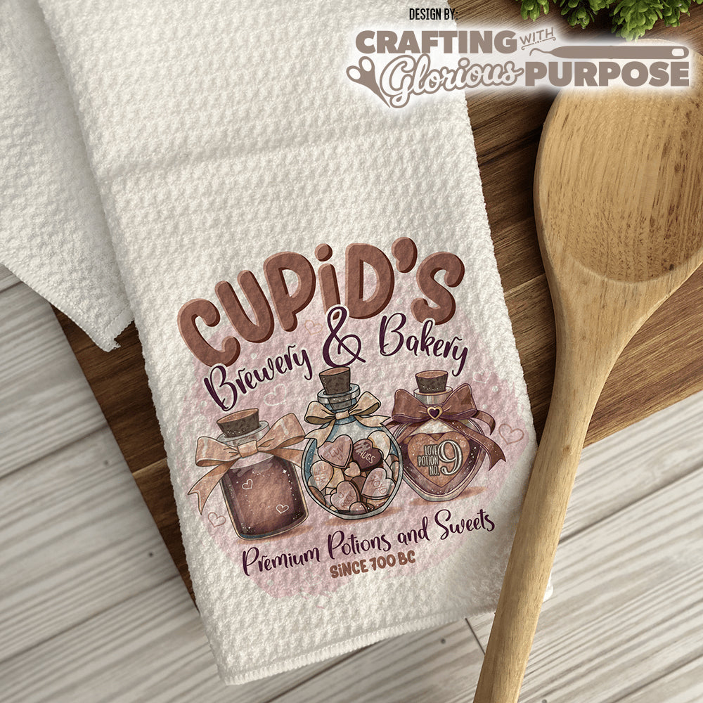 Kitchen towel with 'Cupid's Brewery & Bakery' design on a wooden surface with a wooden spoon.