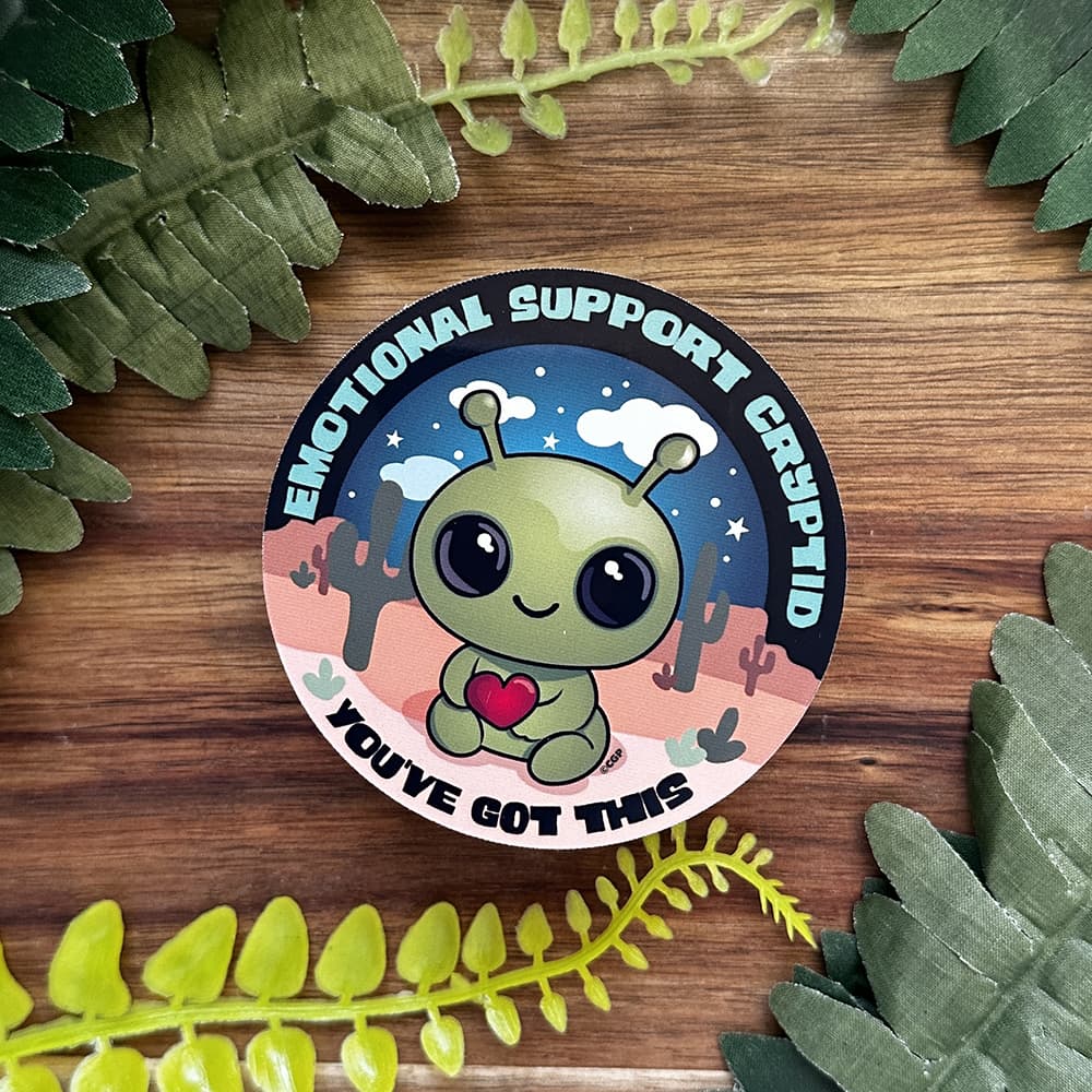Circular sticker with a cartoon alien character surrounded by cacti and stars on a wooden background with green leaves.