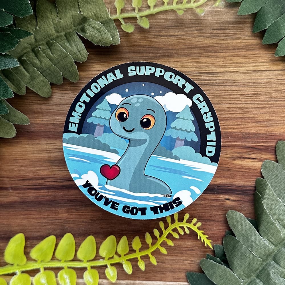 Emotional Support Cryptid sticker with heart and text on a wooden background with leaves