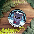 Circular sticker with a cartoon character holding a heart, surrounded by green leaves on a wooden surface.