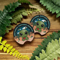 Two 'Emotional Support Crapid' stickers on a wooden surface with green leaves around.
