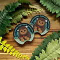 Two stickers with cartoon characters and text on a wooden surface with leaves around.