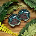 Two Emotional Support Mothman car coasters on a wooden surface with green leaves.