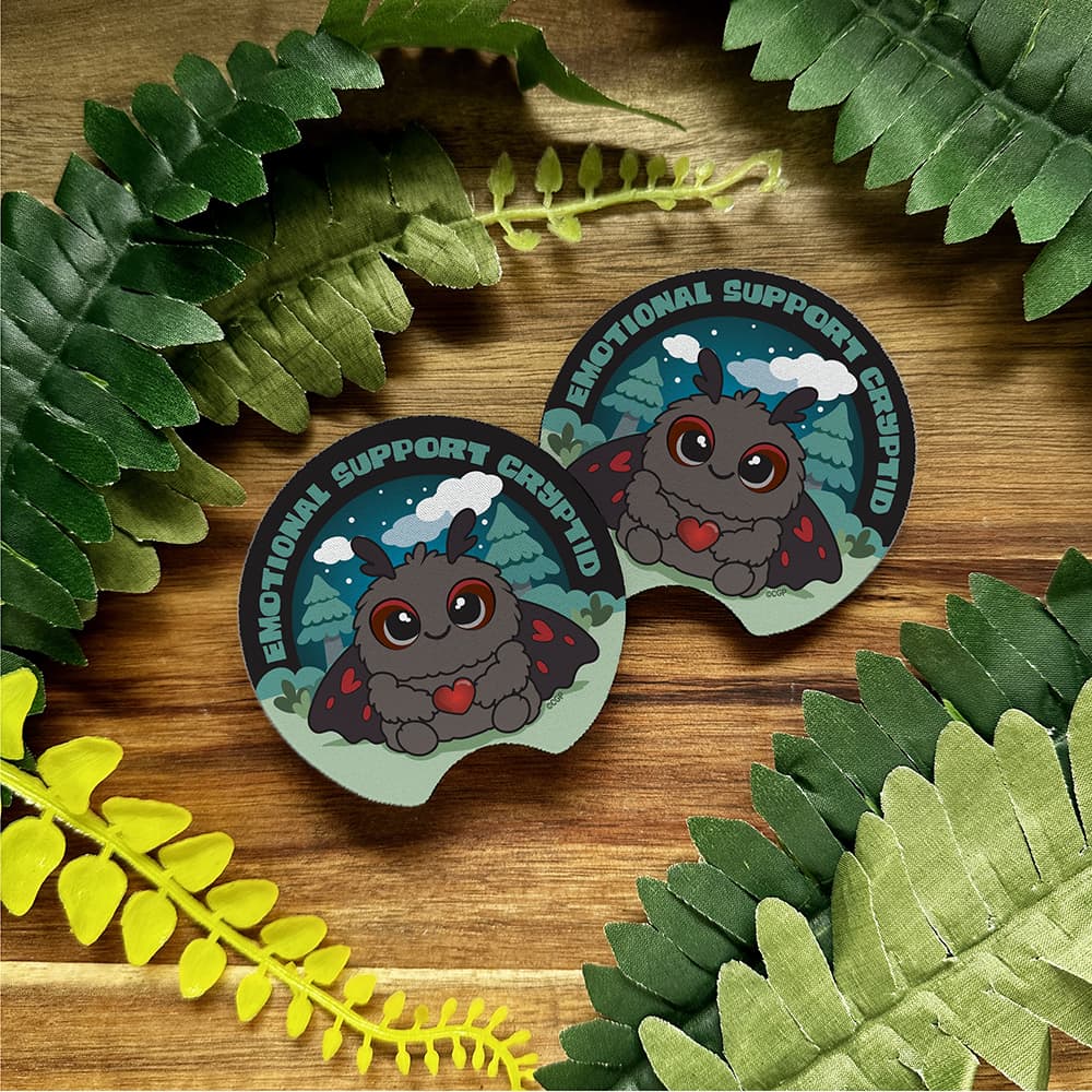 Two Emotional Support Mothman car coasters on a wooden surface with green leaves.