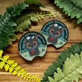 Two 'Emotional Support Cat' themed coasters on a wooden surface with green leaves.