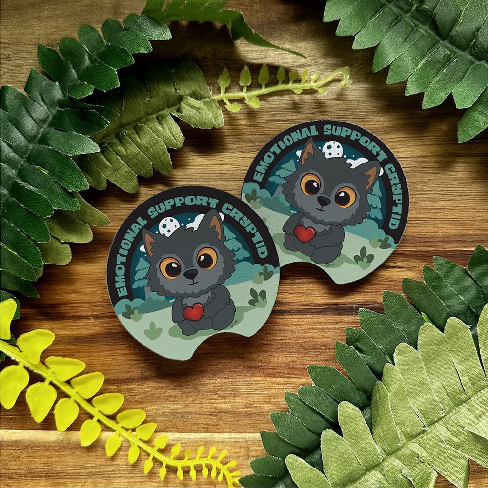 Two 'Emotional Support Cat' themed coasters on a wooden surface with green leaves.