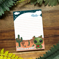 Notepad with a cute alien design on a wooden surface with green leaves.