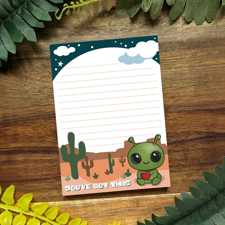 Notepad with a cute alien design on a wooden surface with green leaves.