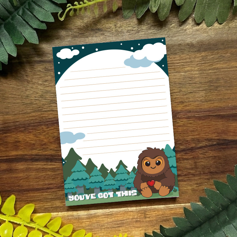 Notebook with cartoon Bigfoot design on a wooden surface with green leaves.