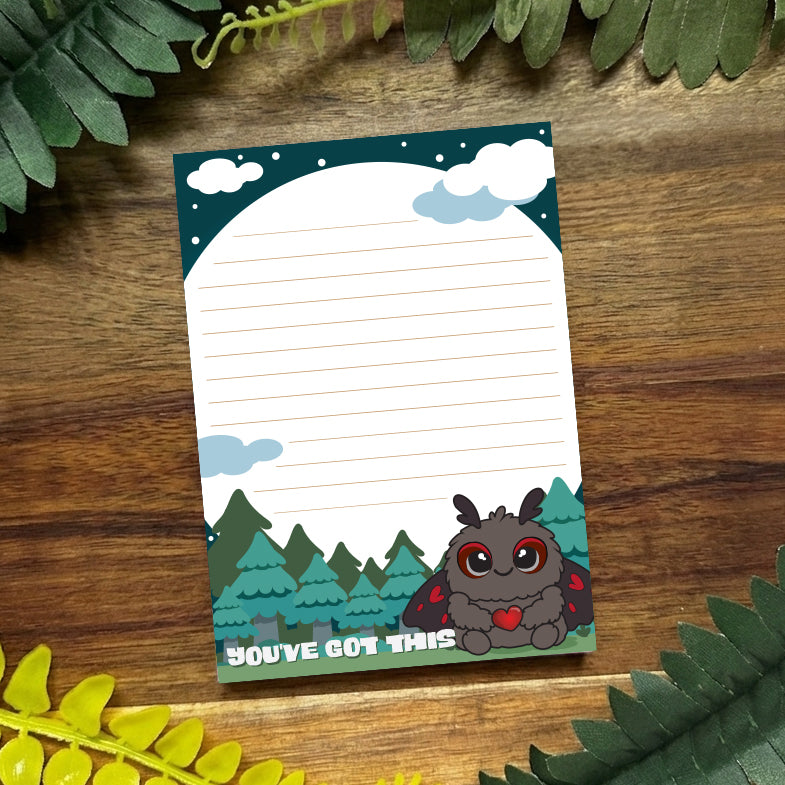 Notebook with a cute mothman design and text 'You've Got This' on a wooden surface with green leaves.