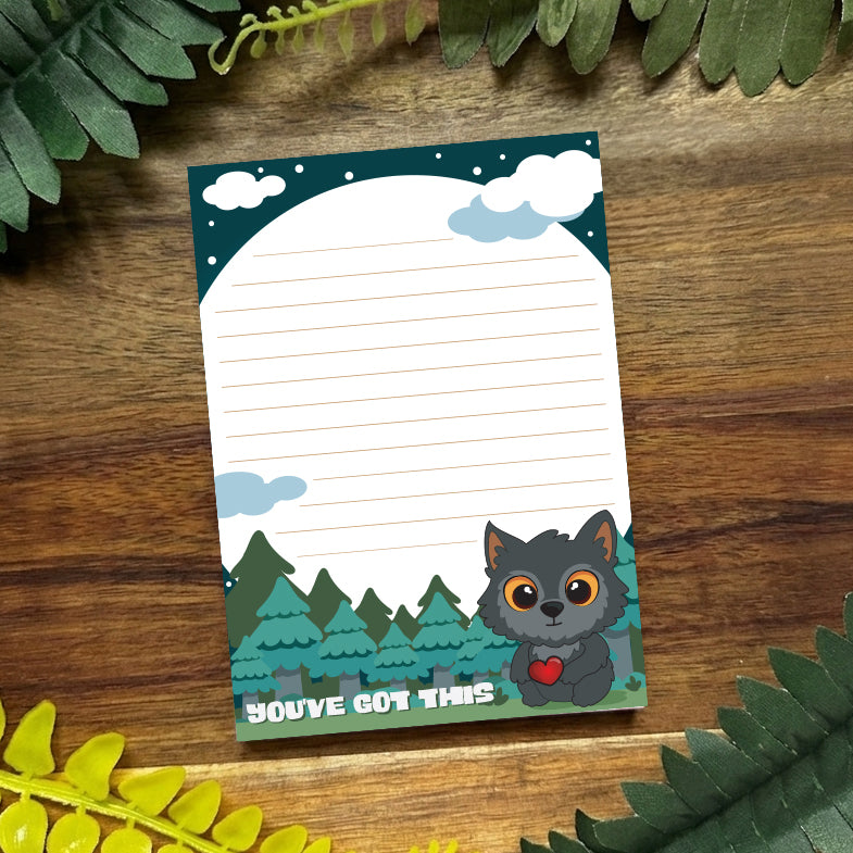Notepad with a cute animal illustration and 'You've Got This' text on a wooden surface with green leaves.
