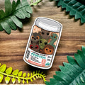 Sticker of a jar labeled 'Emotional Support Cryptids' on a wooden surface with leaves.