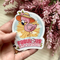 Sticker with a flamingo design and 'Fabulous Like a Flamingo' text held by a hand with pink flowers in the background.