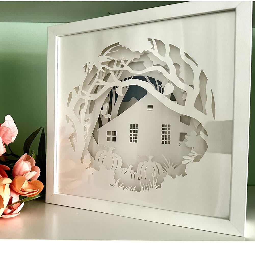 Framed paper cutout of a house and trees with flowers on a table
