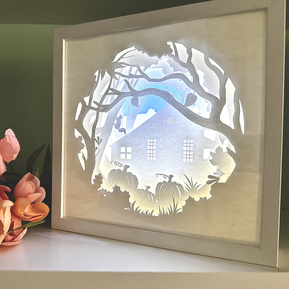 Framed paper cut art of a house and trees with pumpkins on a white surface.