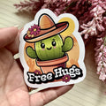 Sticker of a cactus with a hat and 'Free Hugs' text held in a hand with flowers in the background.