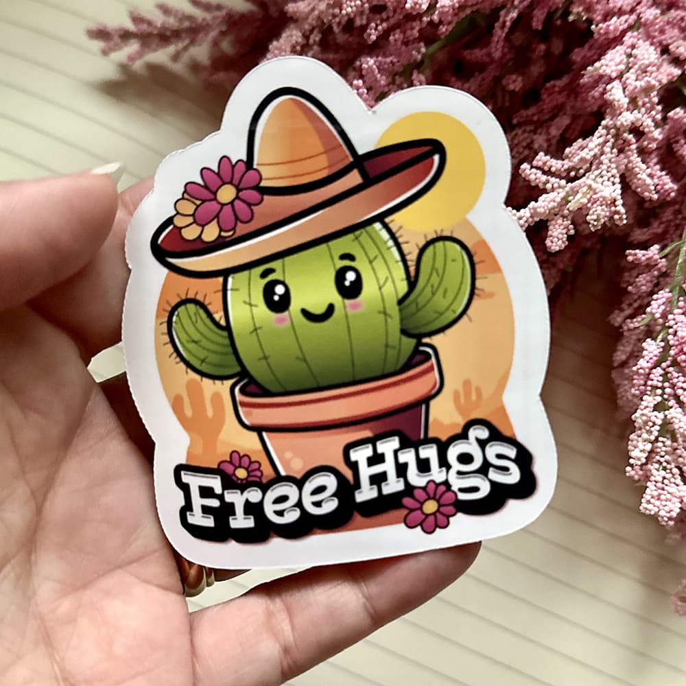 Sticker of a cactus with a hat and 'Free Hugs' text held in a hand with flowers in the background.