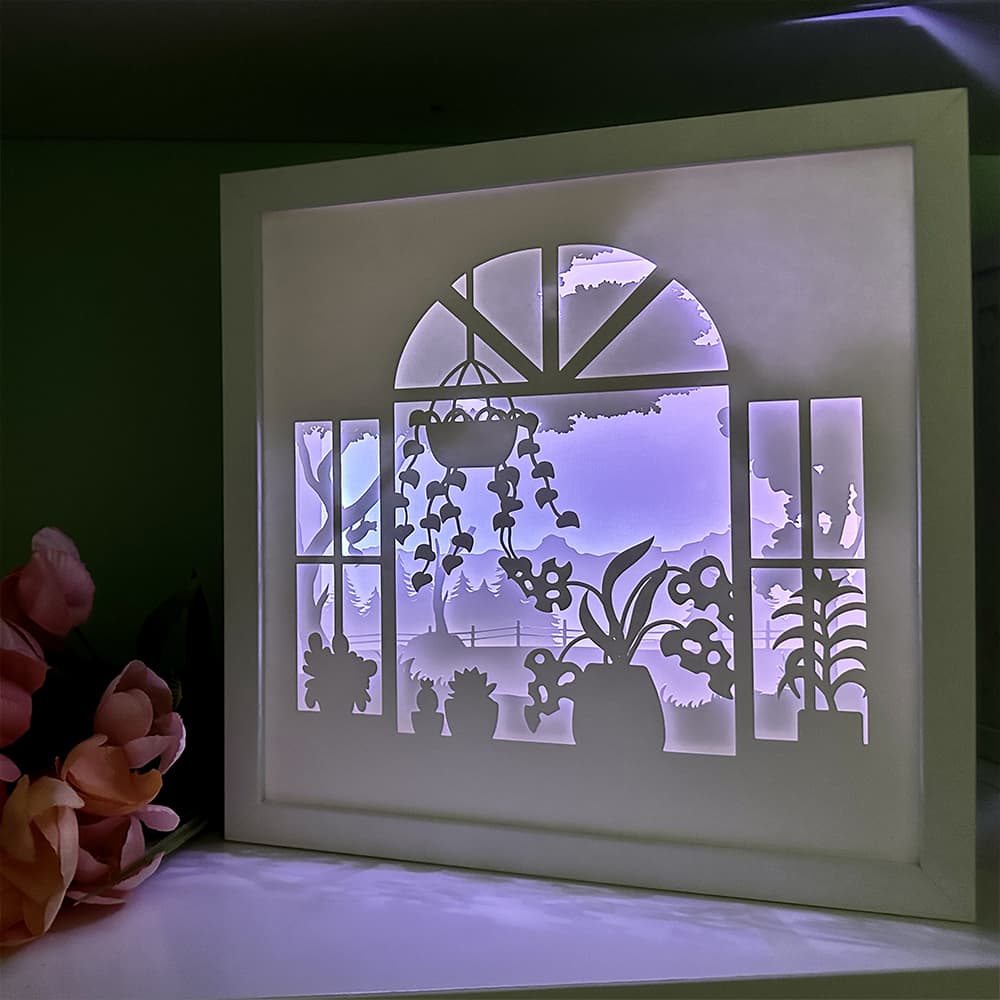 Framed paper cutout art of a garden scene with plants and flowers, illuminated from behind.