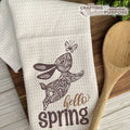 White towel with brown floral design and 'hello spring' text on a wooden surface with a wooden spoon.
