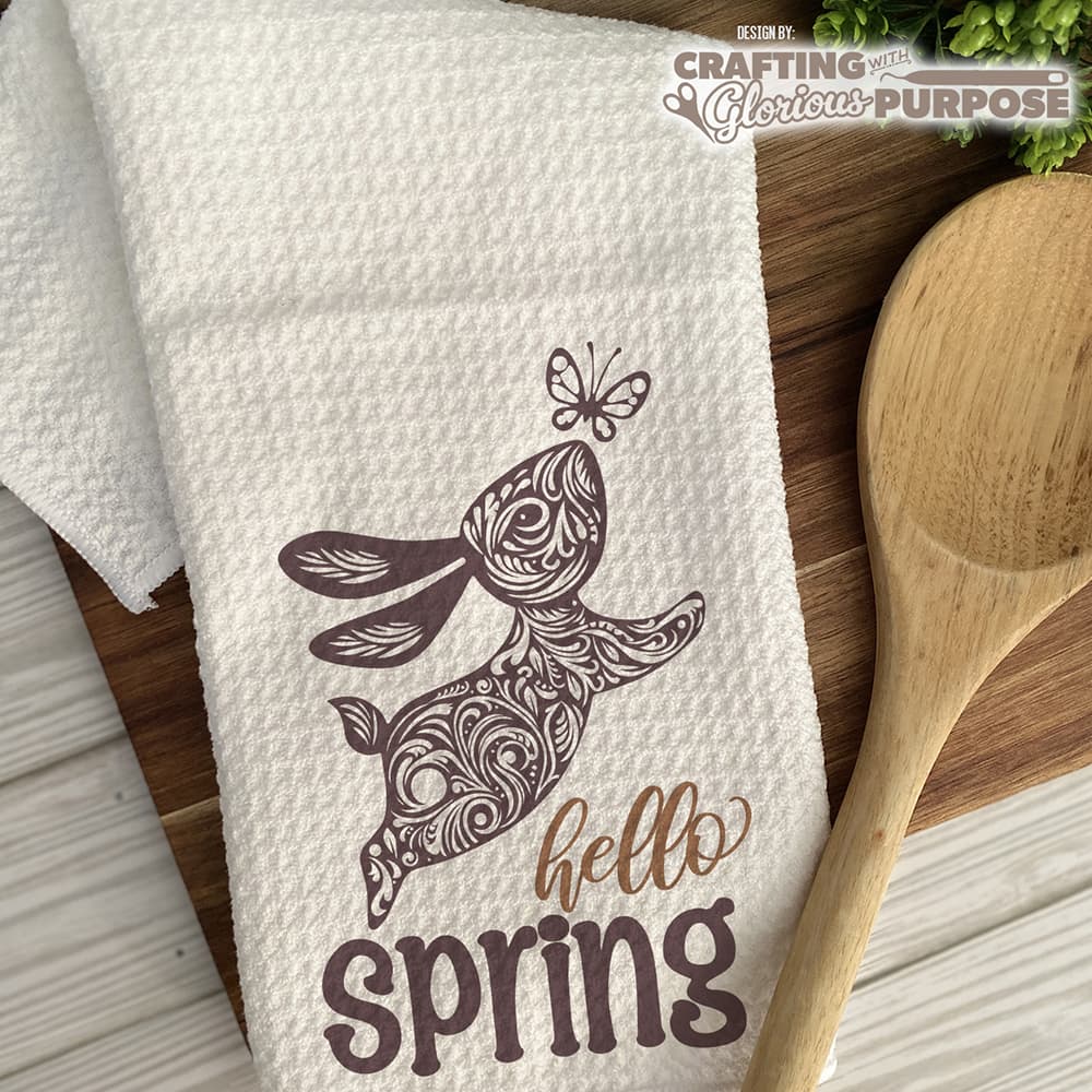White towel with brown floral design and 'hello spring' text on a wooden surface with a wooden spoon.