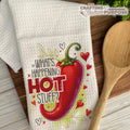 Dish towel with a red pepper graphic and text on a wooden surface with a wooden spoon.
