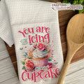 Decorative kitchen towel with a cupcake design and text on a wooden surface.