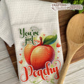  Dish towel with peach illustration and text 'You're just peachy' on a wooden surface.