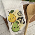 Kitchen towel with cartoon lemons and pears and text, next to a wooden spoon on a wooden surface.