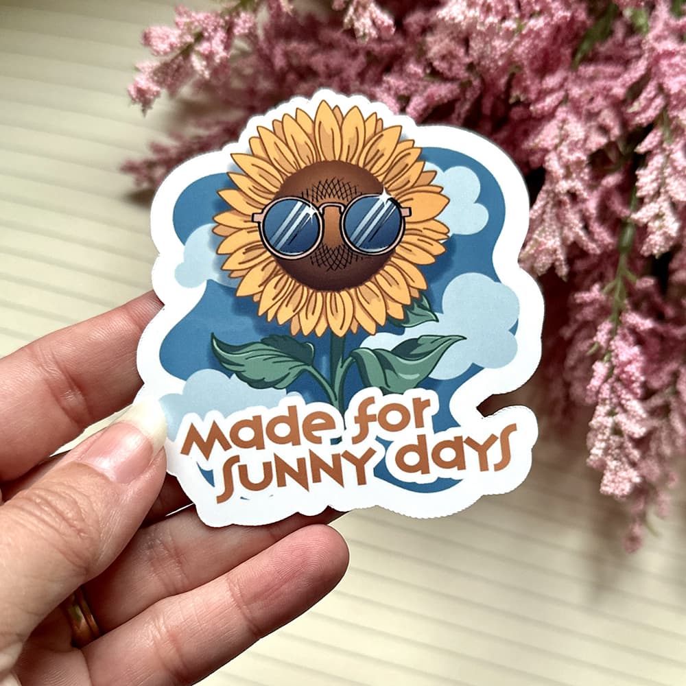 Sticker with a sunflower wearing sunglasses and 'Made for Sunny Days' text, held by a hand with pink flowers in the background.