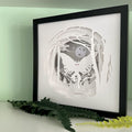 Framed paper cut art of Mothman in a forest with a moonlit sky, placed on a light surface with greenery.