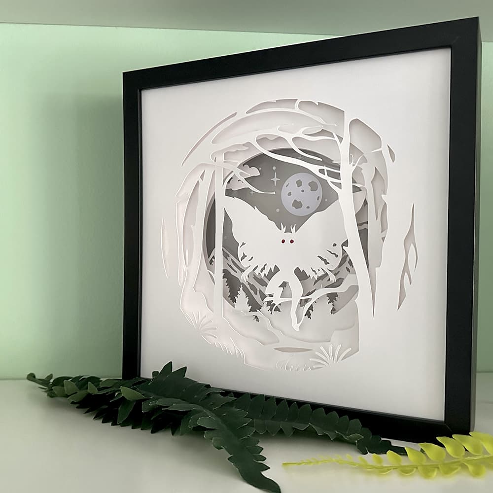 Framed paper cut art of Mothman in a forest with a moonlit sky, placed on a light surface with greenery.