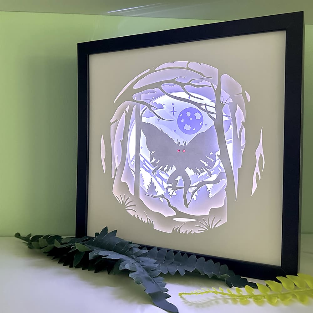 Framed paper cut art of Mothman in a forest with a moonlit sky, placed on a light surface with greenery.