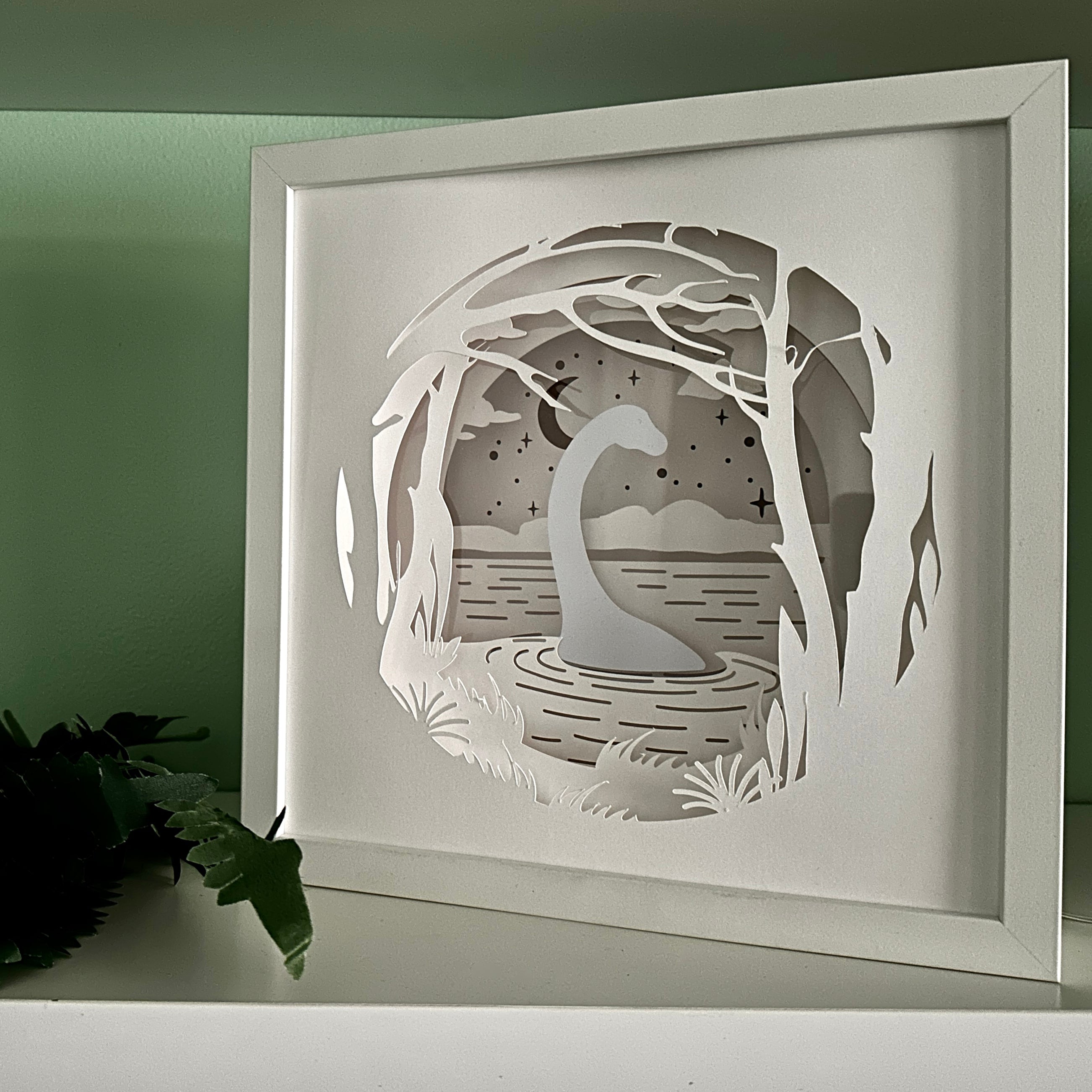Framed paper art of a Nessie by a lake with trees and mountains in the background.
