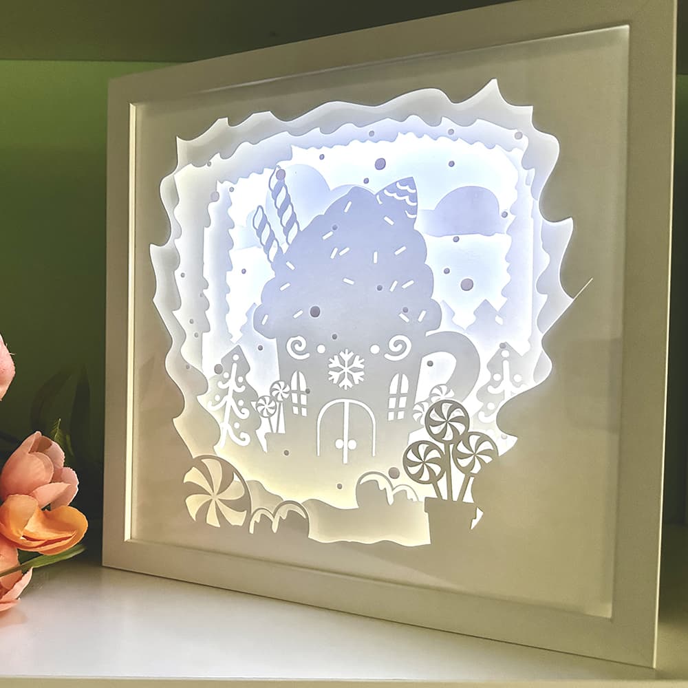 Framed paper cutout art of a gingerbread house with candy cane trees on a white surface.