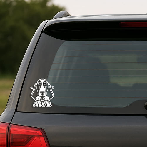 Car window with a sticker of a dog's face and 'Nose Artist on Board' text.