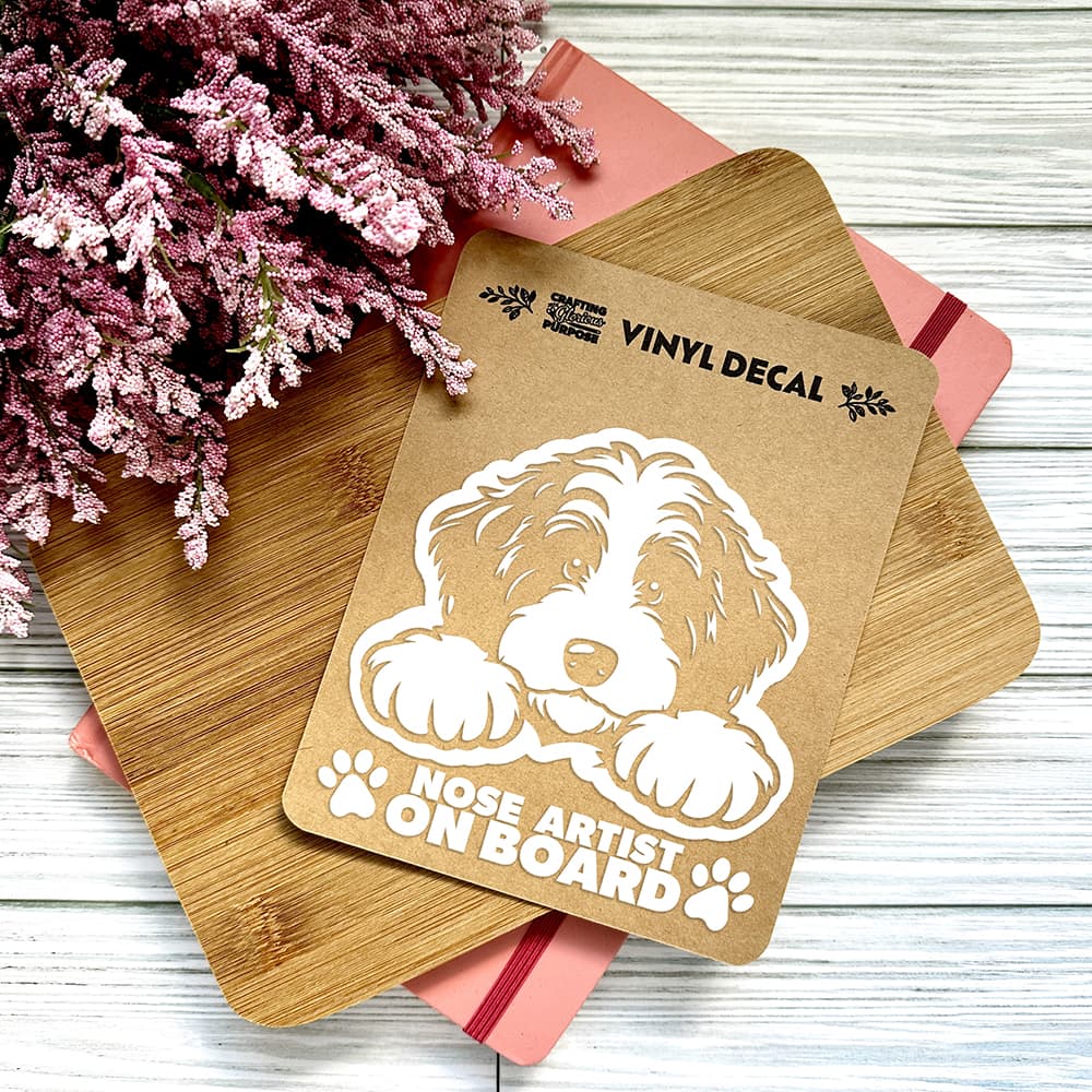 Vinyl decal of a dog with 'Nose Artist on Board' text on a wooden surface with pink flowers.