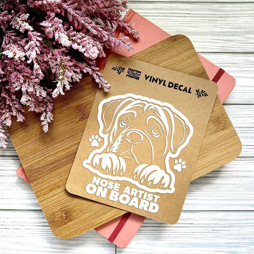 Vinyl decal of a dog with 'Nose Artist On Board' text on a wooden surface with pink flowers.