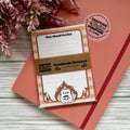 Handmade notepad with cat illustration and text on a wooden surface with pink flowers.