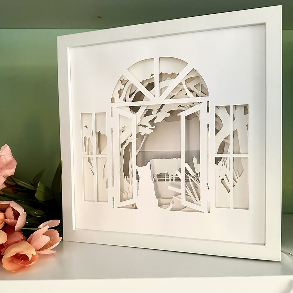 Framed paper art of a cat on a farm landscape with trees on a white surface.