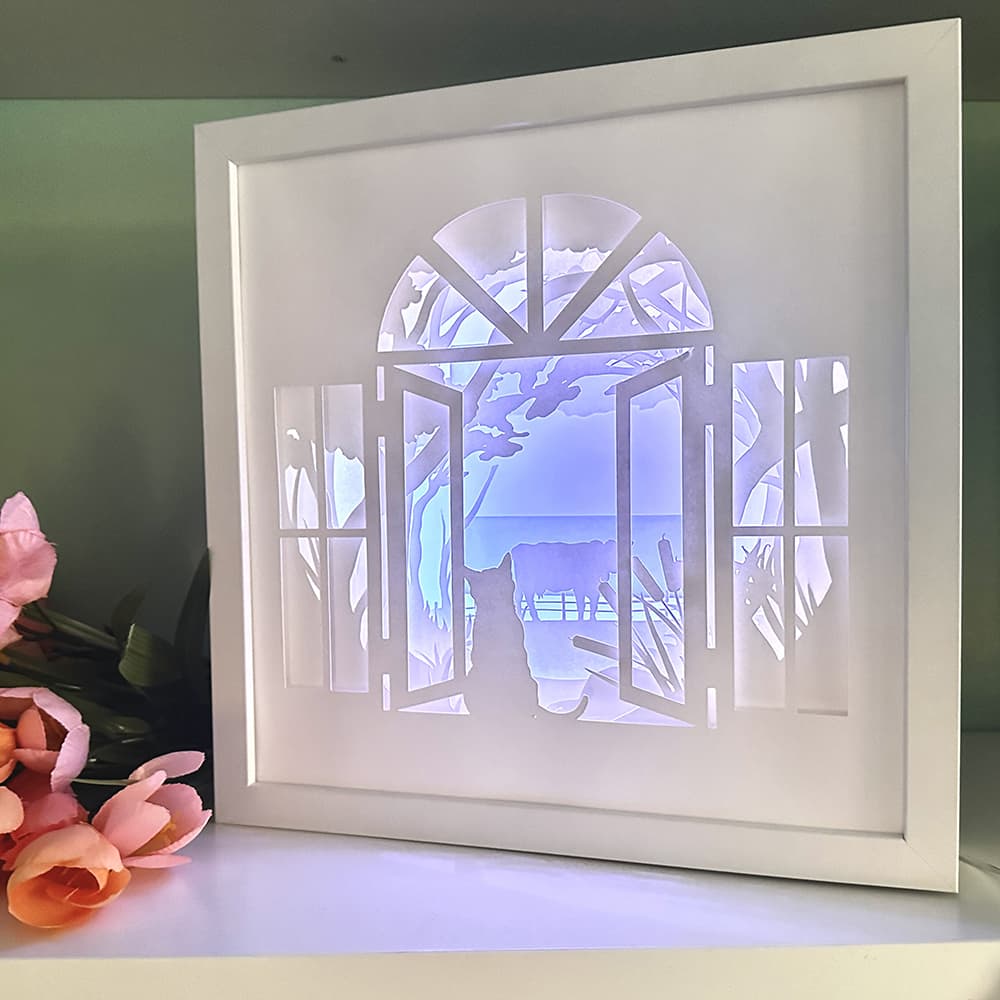 Framed paper cutout of a cat in a window with pink flowers on a table.
