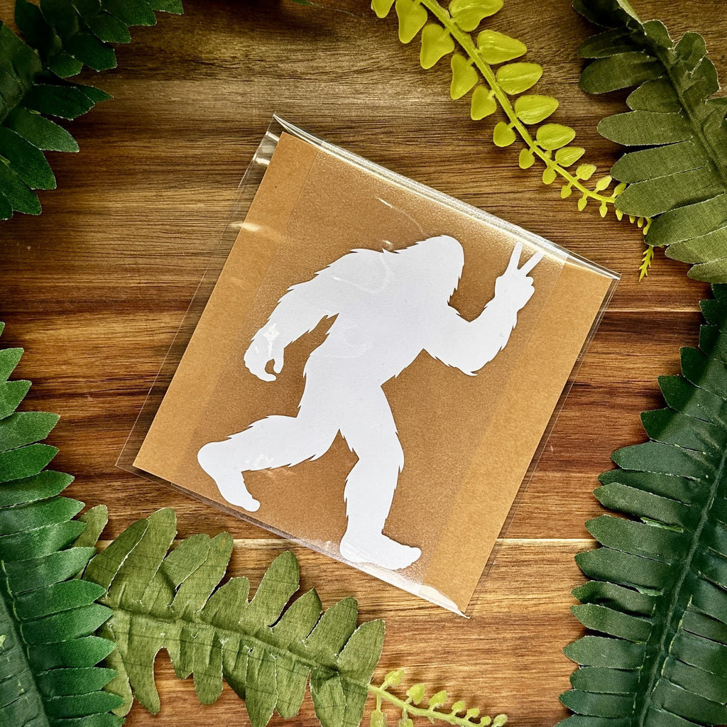 Vinyl decal with white Bigfoot silhouette on a brown paper background, surrounded by green leaves.