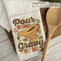 Kitchen towel with 'Pour some gravy on me' design on a wooden surface with a wooden spoon.