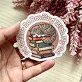 Sticker with a cat and books design held by a hand, with pink flowers in the background.