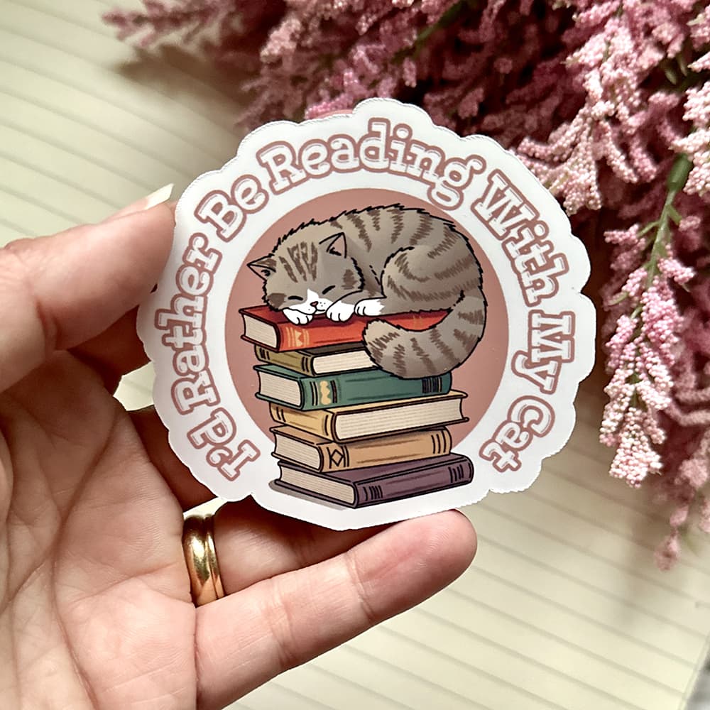 Sticker with a cat and books design held by a hand, with pink flowers in the background.
