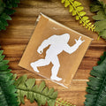 Vinyl decal with a white silhouette of Bigfoot, on a brown paper surrounded by green leaves on a wooden surface.
