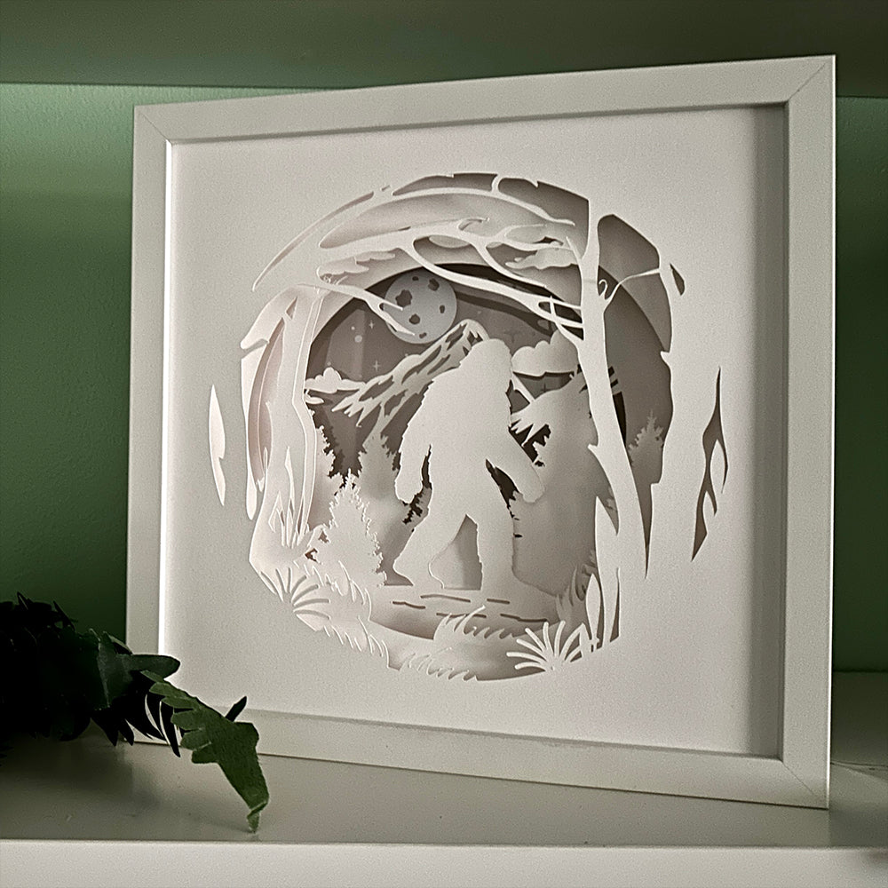 Framed paper cutout of a forest scene with a moon and a silhouette of a bigfoot.