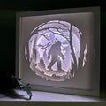 Framed paper cutout of a silhouette of a bigfoot in a forest with a moonlit sky, illuminated by a light source.
